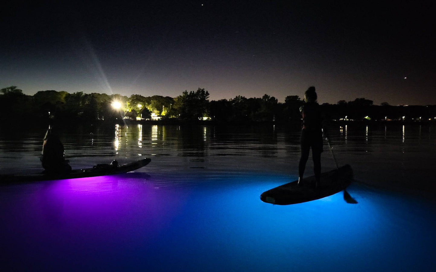 Two friends enjoy a night SUP together with bright paddleboard lights illuminating the water below their SUPs.